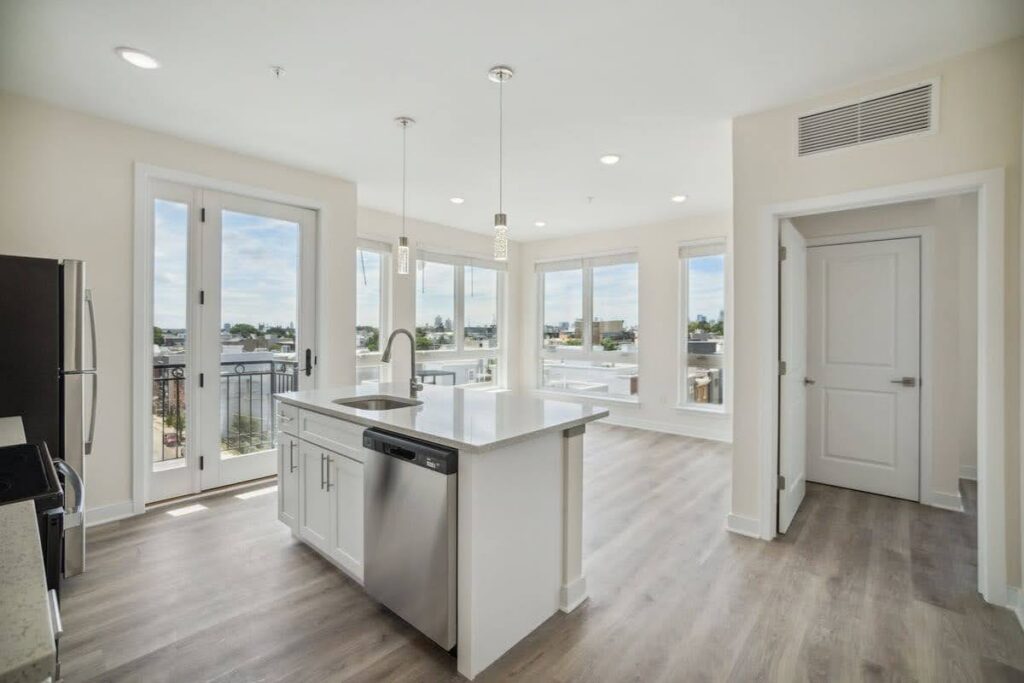 Bright apartment kitchen with white cabinets, island sink, stainless steel appliances, and large windows with balcony access
